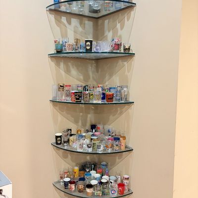 Sale Photo Thumbnail #251: Large collection of novelty, souvenir, and printed shot glasses displayed on a multi-tiered corner glass shelf unit.