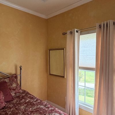 Sale Photo Thumbnail #278: Bedroom featuring a metal bed frame dressed in a burgundy floral comforter, alongside beige grommet curtains hanging over a double-hung window.