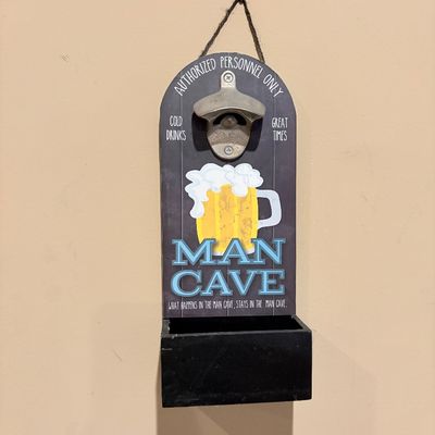 Sale Photo Thumbnail #188: Wall-mounted wooden bottle opener with a 'Man Cave' printed graphic and integrated cap catcher, suspended by a twine hanger.
