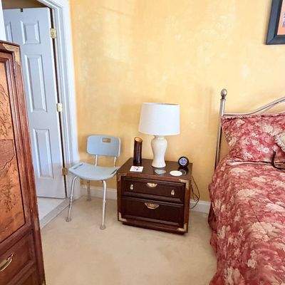 Sale Photo Thumbnail #286: Bedroom corner set with a campaign-style nightstand, cream ceramic table lamp, adjustable medical shower chair, and partial views of a bed and tall armoire.