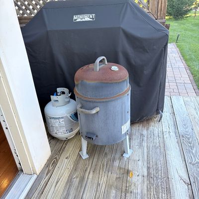 Sale Photo Thumbnail #94: Small vertical bullet-style smoker with rusted lid and temperature gauge, positioned next to a propane tank and a covered Monument brand grill on an outdoor deck.