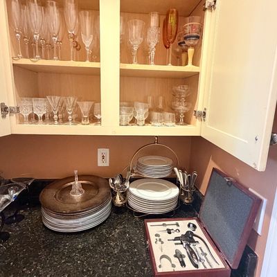 Sale Photo Thumbnail #107: A cabinet filled with various glassware and stemware above a countertop displaying stacked fine china plates, flatware, and a boxed wine tool set.