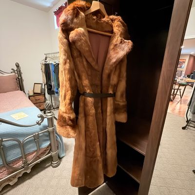 Sale Photo Thumbnail #223: Full-length honey-brown fur coat with wide lapels and a dark leather belt, hanging on a wooden wardrobe door.