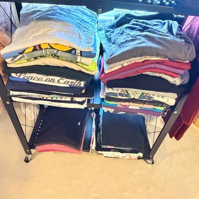 Sale Photo Thumbnail #267: Multiple stacks of folded graphic T-shirts and tops arranged on the shelves of a black metal rolling rack.