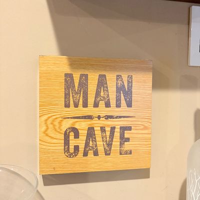 Sale Photo Thumbnail #187: Square wooden novelty sign reading 'MAN CAVE' in a distressed dark gray font with a central decorative motif.