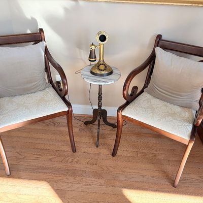 Sale Photo Thumbnail #132: Pair of vintage mahogany armchairs with pattern upholstered seats, paired with a small marble-top accent table displaying an antique-style brass candlestick telephone.