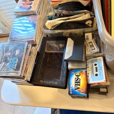 Sale Photo Thumbnail #174: A collection of vintage media including stacks of music CDs, cassette tapes, a Fujifilm VHS-C tape, and a clear plastic storage bin filled with 45 RPM vinyl records.