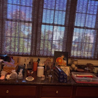Sale Photo Thumbnail #157: An assortment of home decor items, including candles, decorative pottery, framed artwork, and storage baskets, arranged on a dark countertop in front of a window with woven bamboo blinds.