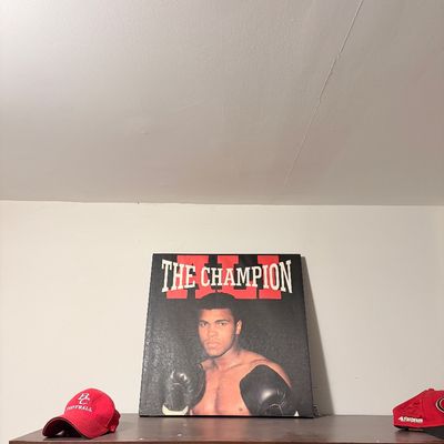 Sale Photo Thumbnail #224: Muhammad Ali 'The Champion' canvas art print displayed on top of a wooden cabinet alongside two baseball caps.