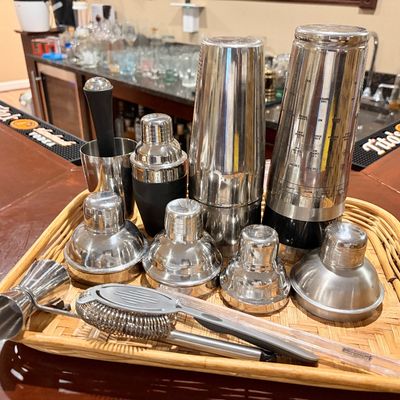 Sale Photo Thumbnail #195: Collection of stainless steel cocktail shakers, strainers, a jigger, and bar tools displayed on a woven wicker tray.