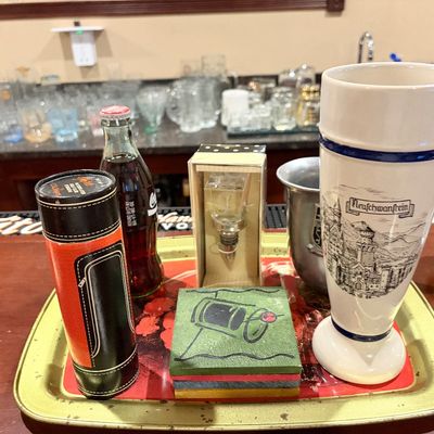 Sale Photo Thumbnail #194: Collection of barware items on a metal tray, including a ceramic Neuschwanstein beer glass, a boxed bottle stopper, coasters, and a unopened glass Coca-Cola bottle.