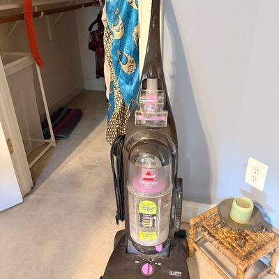 Sale Photo Thumbnail #263: Bissell CleanView Helix Deluxe bagless upright vacuum cleaner with attached power cord and accessories, standing on a carpeted floor next to a small wooden stool.
