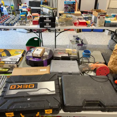 Sale Photo Thumbnail #80: Large collection of power tools, hand tools, hardware cases, and cleaning supplies organized on folding tables, including a Dremel 3000 kit, a Ridgid sander, and a Central Pneumatic finish nailer.