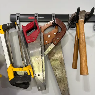 Sale Photo Thumbnail #22: Collection of hand tools hanging on a Rubbermaid FastTrack rail, including handsaws, hacksaws, and wooden-handled claw hammers in used condition.