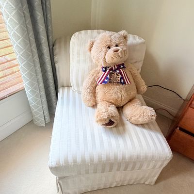 Sale Photo Thumbnail #298: Large beige plush teddy bear wearing an American flag bowtie, sitting on a cream tone-on-tone striped armless slipper chair.