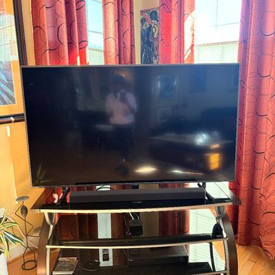 Sale Photo Thumbnail #146: Large flat-screen television with soundbar sitting on a modern curved glass and metal TV stand, featuring lower shelves with electronic components.