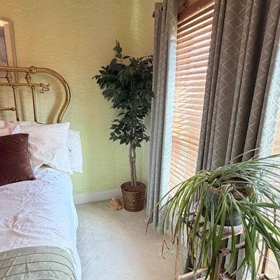 Sale Photo Thumbnail #291: Artificial potted ficus tree sitting in the corner of a bedroom alongside window drapes and a wooden plant stand.