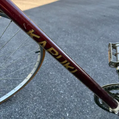 Sale Photo Thumbnail #50: Close-up of a vintage Kapuk bicycle frame in maroon with gold lettering, showing wear consistent with age.