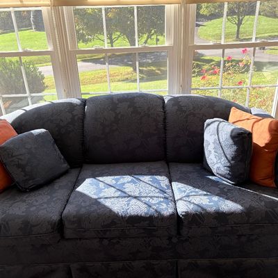 Sale Photo Thumbnail #137: Blue damask patterned upholstered sofa with matching lumbar pillows and contrasting orange accent pillows, set in front of a sunlit bay window.
