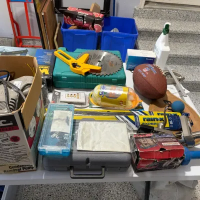 Sale Photo Thumbnail #82: Assortment of household goods, tools, and hardware displayed on a folding table, including a Flotec condensate removal pump, Wilson football, saw blade, and cleaning supplies.