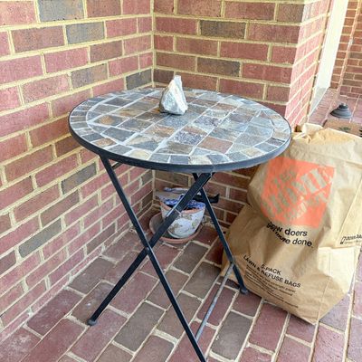Sale Photo Thumbnail #162: Round outdoor bistro table with a slate tile mosaic top and a black folding metal base, shown on a brick porch.