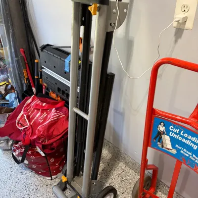 Sale Photo Thumbnail #27: Portable material lifter and hand truck with roller top, alongside a red steel hand truck in a workshop setting.