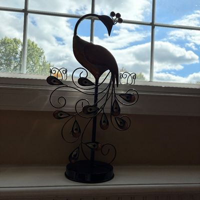 Sale Photo Thumbnail #212: Decorative metal peacock candle holder featuring intricate scrollwork tail feathers and glass votive cups, displayed in front of a window.