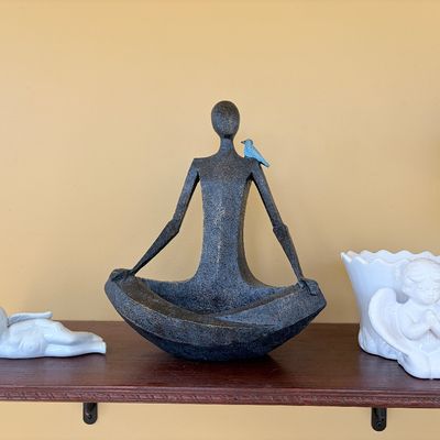 Sale Photo Thumbnail #123: Stylized textured bronze-look figure in a yoga posture with a blue bird perched on its shoulder, displayed alongside two white ceramic cherub figurines on a wooden shelf.