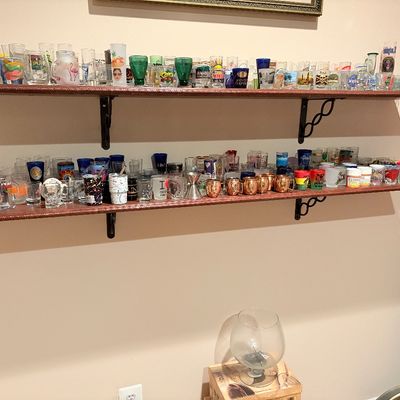 Sale Photo Thumbnail #250: Large collection of novelty shot glasses displayed on two wall-mounted wooden shelves, including various souvenir, printed, and colorful glassware.