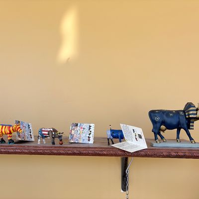 Sale Photo Thumbnail #125: Collection of decorative CowParade cow figurines and an elephant figurine lined up on a wooden shelf, including Egyptian and patriotic themed designs.