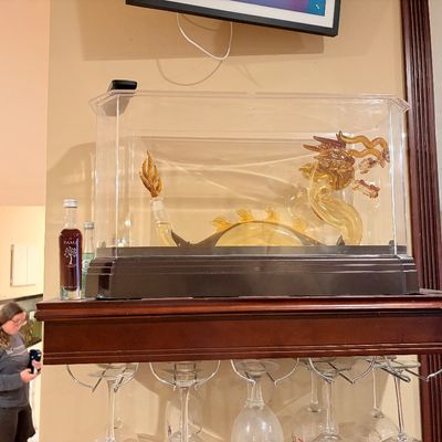 Sale Photo Thumbnail #192: Ornate glass dragon-shaped decanter housed inside a clear acrylic display case with a black base, resting above a wine glass rack.