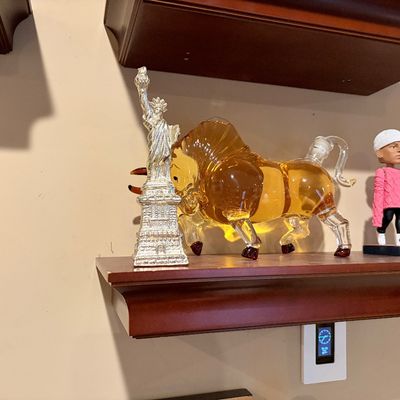 Sale Photo Thumbnail #189: Novelty collectibles displayed on a wooden shelf, including a clear glass charging bull decanter with amber fluid, a silver-toned Statue of Liberty figurine, and a Kyle Kuzma bobblehead.