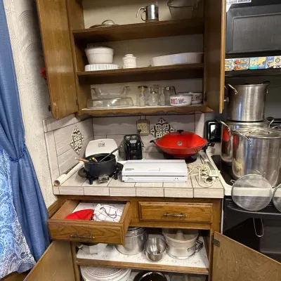 Sale Photo Thumbnail #34: Open wooden kitchen cabinets and countertop filled with various cookware, including stainless steel stock pots, ceramic baking dishes, a vacuum sealer, and small kitchen appliances.