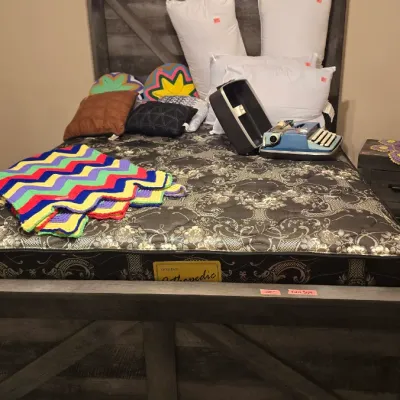 Sale Photo Thumbnail #17: Rustic weathered grey wood full-size bed frame with mattress, assorted throw pillows, a colorful crochet blanket, and a vintage blue portable typewriter.