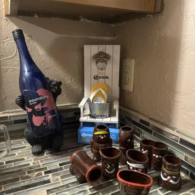 Sale Photo Thumbnail #48: Novelty barware collection featuring vintage ceramic boot and character shot glasses, a Corona Extra chair-shaped wall bottle opener, and a black bear bottle holder.