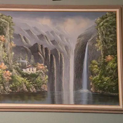 Sale Photo Thumbnail #12: Framed oil painting depicting a dramatic mountain landscape with a waterfall, river, and a small cottage nestled among lush greenery.