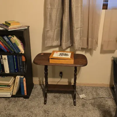 Sale Photo Thumbnail #18: Vintage dark wood two-tier side table with turned legs, featuring a framed Mickey Mouse art piece on top and situated next to a dark bookcase filled with books.