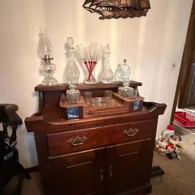 Sale Photo Thumbnail #36: Vintage dark wood dry sink cabinet topped with assorted glassware, including crystal decanters, stemware, and an oil lamp, with a metal ship wall hanging mounted above.
