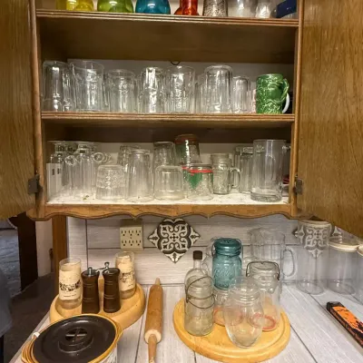 Sale Photo Thumbnail #30: Collection of vintage glassware, including colorful decanters and tumblers arranged in a wooden cabinet, above kitchen accessories like a potpourri crock and glass mason jars.