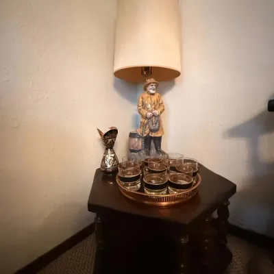 Sale Photo Thumbnail #32: Vintage table lamp featuring a fisherman figural base alongside a tray of lowball glasses and a small metallic vase on a hexagonal wooden end table.
