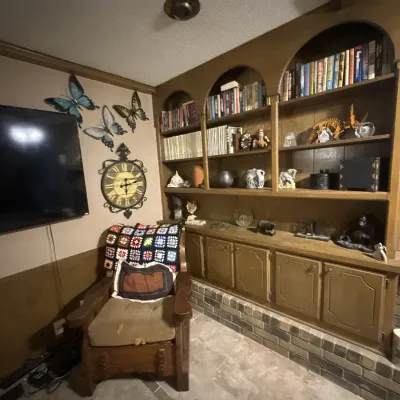 Sale Photo Thumbnail #50: Living room setup featuring a large built-in wooden wall unit with arched shelving, a wooden armchair draped with a colorful crochet blanket, decorative wall clocks, butterfly art, and various knick-knacks.
