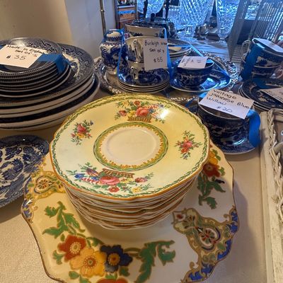 Sale Photo Thumbnail #111: A table set with vintage china and glassware, featuring yellow floral hand-painted plates stacked on a matching serving dish alongside stacks of blue and white Blue Willow teacups, saucers, and dinner plates.