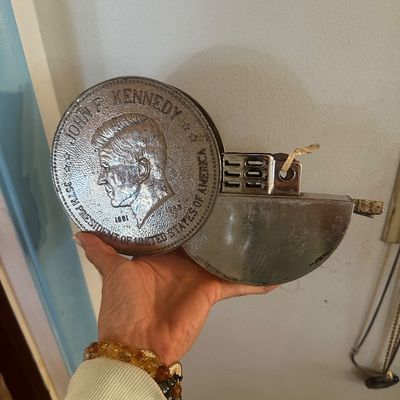 Sale Photo Thumbnail #83: Oversized John F. Kennedy commemorative coin-form table lighter featuring a raised profile portrait and textured metal finish.