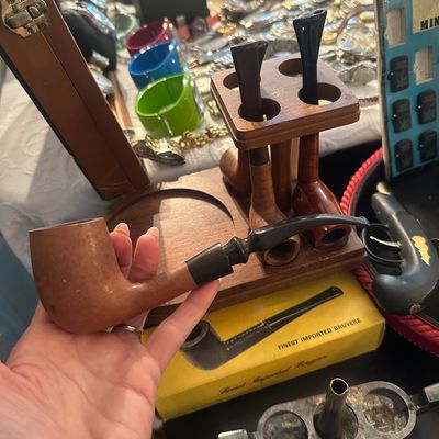 Sale Photo Thumbnail #81: Hand holding a classic wooden smoking pipe alongside a wooden multi-pipe rack display, accompanied by additional tobacciana items and original yellow box.