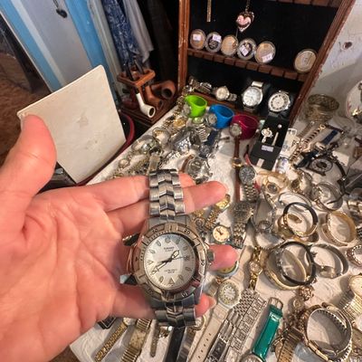Sale Photo Thumbnail #65: Hand holding a stainless steel Casio Quartz 100M wrist watch over a tabletop filled with various vintage and modern watches and jewelry.