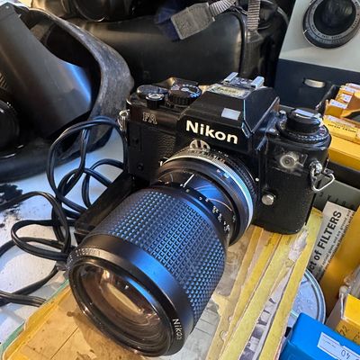 Sale Photo Thumbnail #86: Vintage black Nikon FA 35mm SLR camera fitted with a Nikkor lens, surrounded by various camera equipment.
