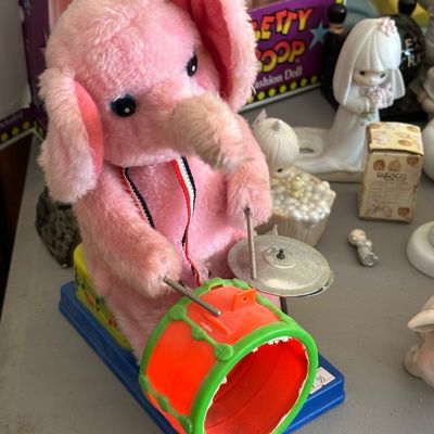 Sale Photo Thumbnail #54: Vintage battery-operated wind-up style mechanical pink plush elephant playing drums, featuring a plastic drum set mounted on a blue base.