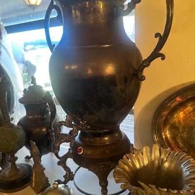Sale Photo Thumbnail #93: Vintage electric silver-plated samovar beverage dispenser with ornate handles and spigot, shown alongside metal accessories and hollowware.