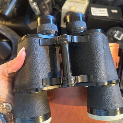 Sale Photo Thumbnail #87: Vintage black leatherette-wrapped binoculars featuring adjustable focus rings, held over a brown leather carrying case.
