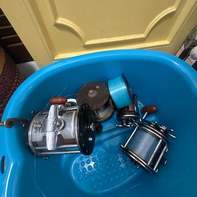 Sale Photo Thumbnail #69: Collection of vintage conventional and trolling fishing reels, including metal and composite models with attached monofilament line, stored in a blue plastic container.